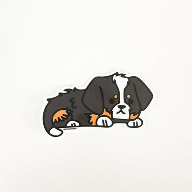 An illustrated bernedoodle puppy sticker with black, tan and white markings laying down